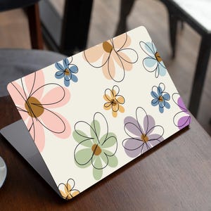 May include: Laptop featuring a floral pattern. The laptop's cover displays a cream backdrop adorned with large, colourful flowers in pink, blue, green, yellow, and purple, outlined in black. The laptop rests on a wooden table.