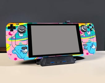 May include: A handheld gaming console featuring a pink skin with a colorful space-themed pattern of blue bears, yellow planets, and rings. The device is docked in a black stand with two USB ports and a central USB-C port, displaying a blank screen.