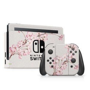 Op de afbeelding: Een witte Nintendo Switch console en controller met een bloemenontwerp met roze kersenbloesems op een lichtgrijze achtergrond. De console heeft het Nintendo Switch logo en de tekst "NINTENDO SWITCH" erop gedrukt.