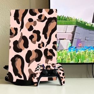 May include: A PlayStation 5 console and controller skin set featuring a pink background with a brown and black leopard print pattern. The controller has black buttons and a white face.