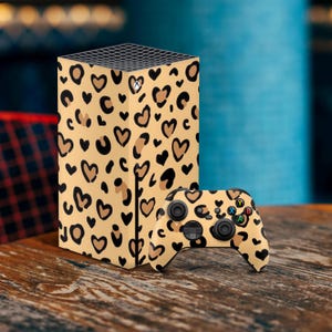 Può includere: Una pelle per console e controller di gioco con stampa leopardata beige e marrone. La console è una scatola rettangolare con bordi arrotondati. Il controller ha un design bianco e nero con pulsanti e un joystick.