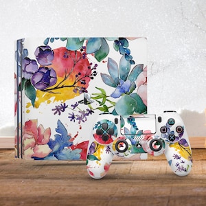 May include: A white game console and matching wireless controller featuring a vibrant watercolor print of purple flowers, blue succulents, and splashes of yellow and red paint. The console displays the PlayStation logo in silver on the front panel.