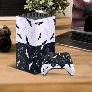 May include: A gaming console and controller set with a bat design. The console has a black and white bat pattern, with the controller mirroring the design. The bat silhouettes are in black and white, creating a Halloween theme. The set is on a wooden surface.