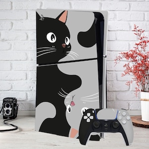 Peut inclure: Une console PlayStation 5 noire et grise avec une manette assortie. La console est ornée d'un motif de deux chats de dessin animé, l'un noir et l'autre gris, sur fond gris. La manette est noire et grise.