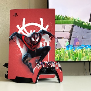 May include: A red and black PlayStation 5 console with a custom skin featuring a stylized image of Spider-Man in a red and black suit. The console is on a white surface with a matching red and black PlayStation 5 controller.