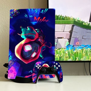 May include: A black and blue PlayStation 5 console with a custom skin featuring a cartoon character from the movie Spider-Man: Into the Spider-Verse. The skin features the character Miles Morales in a red and blue suit with a black spider design. The text "It is all Miles" is visible on the skin. A black and white PlayStation 5 controller with a matching skin is also visible in the image.