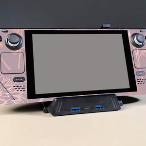 Può includere: Console di gioco portatile con un design floreale nei toni del rosa e del rame. La console ha un ampio schermo ed è collegata a una docking station nera con porte USB. La parola "STEAM" è visibile sulla console.