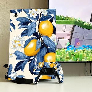 Könnte beinhalten: Eine weiße PlayStation 5 Konsole mit einem blauen und gelben Blumendesign mit Zitronen und weißen Blüten. Die Konsole steht neben einem passenden PlayStation 5 Controller.