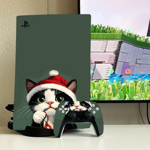 Puede incluir: Una piel de consola PlayStation 5 verde y una piel de mando a juego con un gato blanco y negro con un gorro de Papá Noel. El gato sostiene una guirnalda de luces. La consola PlayStation 5 está sobre una superficie blanca.