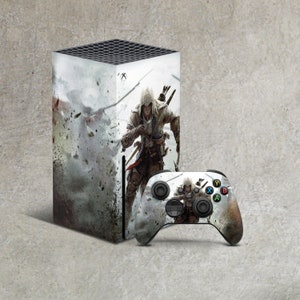 May include: A white and gray video game console with a design featuring a character in a hooded cloak and a sword. The console is next to a black video game controller with the same design.