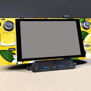 Può includere: Una console di gioco portatile con una skin gialla a motivi di limoni e foglie verdi, dotata di joystick, pulsanti e pad direzionale neri, posizionata su una docking station nera con due porte USB blu e una porta centrale.