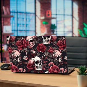 May include: A laptop with a black cover featuring a floral and skull design. The design includes red roses and pink skulls on a black background. The laptop is open on a wooden desk with a mouse and a small potted plant.