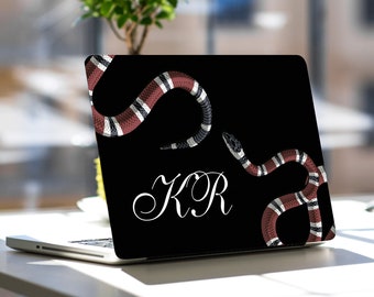 May include: Black laptop skin with a design of two snakes in red, white, and black. The laptop skin has the initials "KR" in white script. The laptop is open on a white surface.