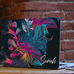 May include: A laptop with a black background and a colorful floral design. The design features pink, blue, and yellow flowers with black outlines. The name "Sarah" is printed in white on the bottom right corner of the laptop.