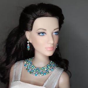 May include: A doll with long, dark hair and blue eyes, wearing a sparkling necklace and matching earrings. The necklace and earrings are adorned with colorful, iridescent gemstones. The doll is dressed in a white dress.