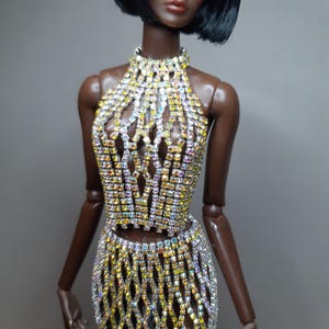 May include: A two-piece doll outfit consisting of a sleeveless halter-neck top and a matching skirt, both constructed from a lattice of iridescent, multi-colored rhinestones that catch the light, creating a shimmering, grid-like appearance.