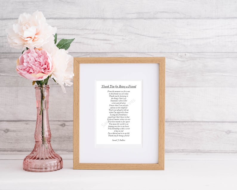 Op de afbeelding: Een ingelijst afdruk met een gedicht met de titel "Thank You for Being a Friend". Het gedicht is gedrukt in zwarte inkt op wit papier. De lijst is van lichtbruin hout met een witte passe-partout. De afdruk ligt op een licht houten oppervlak met roze bloemen in een vaas.