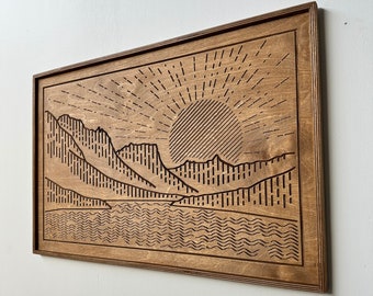 Puede incluir: Arte de pared de madera con un diseño de paisaje cortado con láser. La obra de arte representa una cordillera, un sol y una masa de agua. El marco es de color marrón oscuro y la obra de arte es de color marrón claro.