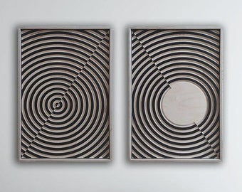 May include: Two black and white abstract wall art pieces with a circular design. The pieces are framed in a thin, silver frame and are hanging on a white wall above a beige sofa.