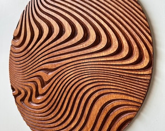 May include: A round, wooden wall art piece with a carved, wave-like pattern. The artwork is a warm brown color and has a three-dimensional effect due to the carved grooves. The piece is mounted on a white wall.