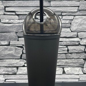 May include: A black reusable tumbler with a clear dome lid and a black straw. The tumbler is made of a matte black material. The lid has a hole for the straw. The tumbler is sitting on a black base.