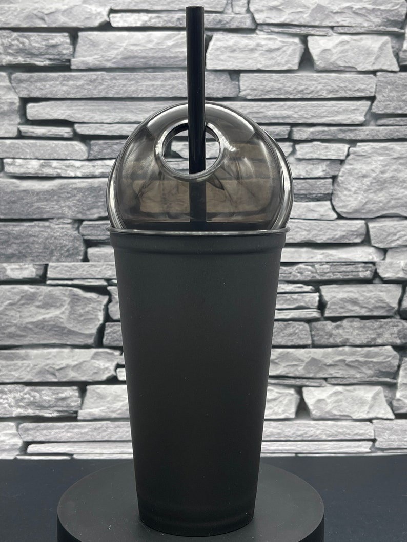 May include: A black reusable tumbler with a clear dome lid and a black straw. The tumbler is made of a matte black material. The lid has a hole for the straw. The tumbler is sitting on a black base.