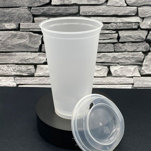 May include: A clear, frosted plastic cup with a matching lid. The lid has a small opening for a straw. The cup and lid are on a black surface against a grey stone background. The cup is designed for cold drinks.