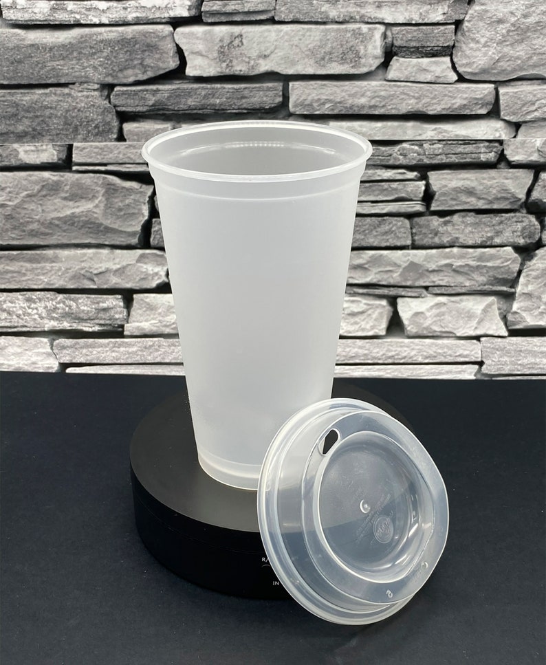 May include: A clear, frosted plastic cup with a matching lid. The lid has a small opening for a straw. The cup and lid are on a black surface against a grey stone background. The cup is designed for cold drinks.