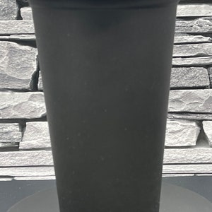May include: A black travel mug with a black lid. The mug is cylindrical with a slightly tapered design. The lid is a snap-on style. The mug is sitting on a black surface.