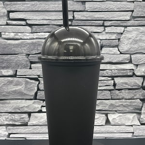 May include: A black plastic tumbler with a clear dome lid and a black straw. The cup is cylindrical with a slightly tapered shape. The lid is clear, allowing a view of the contents. The straw is inserted through a hole in the lid.