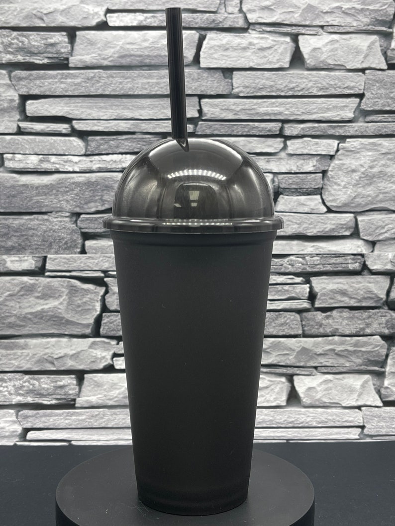 May include: A black plastic tumbler with a clear dome lid and a black straw. The cup is cylindrical with a slightly tapered shape. The lid is clear, allowing a view of the contents. The straw is inserted through a hole in the lid.