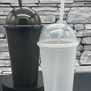 May include: Two plastic tumblers with dome lids and straws. One tumbler is black, the other is translucent white. Both have a tapered shape and are designed for cold beverages. The straws are black and white.