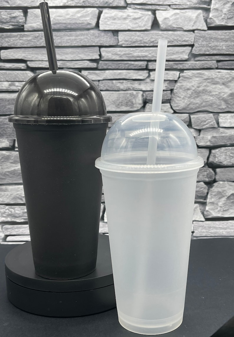 May include: Two plastic tumblers with dome lids and straws. One tumbler is black, the other is translucent white. Both have a tapered shape and are designed for cold beverages. The straws are black and white.