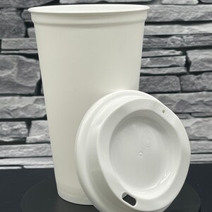 May include: A white paper coffee cup and lid. The cup is tall and conical, with a rolled edge. The lid is placed beside the cup, showing the drinking aperture. The cup and lid are on a black surface, against a grey stone wall.