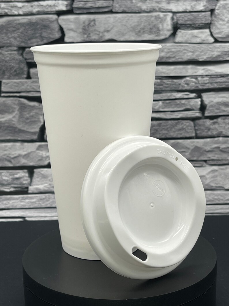 May include: A white paper coffee cup and lid. The cup is tall and conical, with a rolled edge. The lid is placed beside the cup, showing the drinking aperture. The cup and lid are on a black surface, against a grey stone wall.