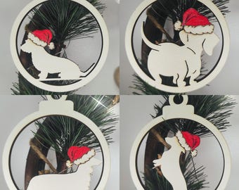 May include: Four white wooden Christmas ornaments shaped like a ball with a dachshund dog wearing a Santa hat inside each ornament. The ornaments are hanging from a Christmas tree.