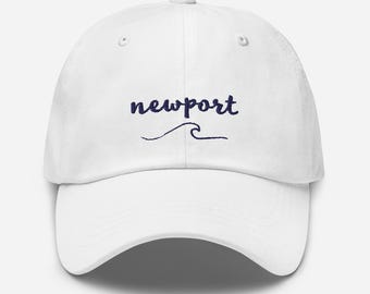 May include: White baseball cap with the word "newport" in navy blue script, with a navy blue wave graphic underneath. The cap has a curved brim and an adjustable strap.