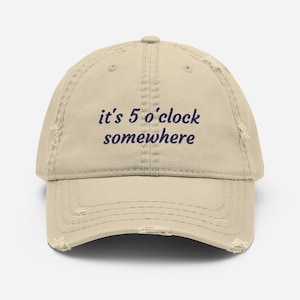 May include: A distressed beige baseball cap with the embroidered phrase "it's 5 o'clock somewhere" in navy blue. The cap has a curved brim and a worn, vintage appearance.
