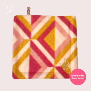 May include: A double-sided skate towel with a geometric pattern in shades of pink, yellow, and white. The towel has a small glittery loop for hanging and a gold embroidered initial. The text "Double-sided Skate towel" is on a pink circle.