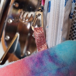May include: A close-up of a pink and blue tie-dye fabric bag with a silver zipper and a pink glitter zipper pull.