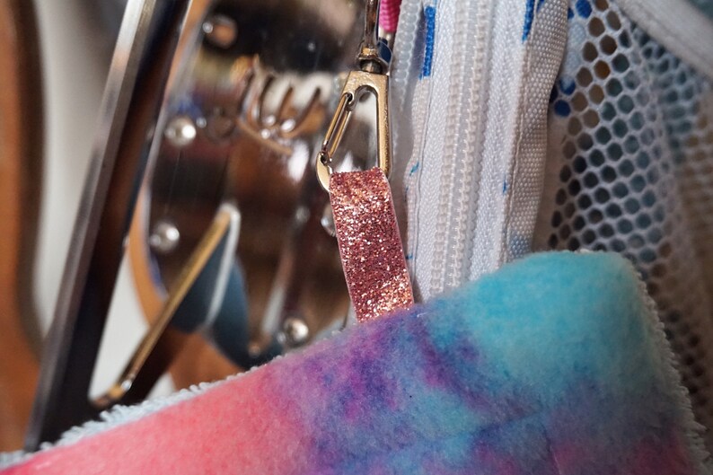 May include: A close-up of a pink and blue tie-dye fabric bag with a silver zipper and a pink glitter zipper pull.