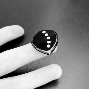 May include: A black ring with a smooth, polished finish and a teardrop-shaped face. The surface of the ring features a curved row of five small, circular white inlays arranged in a vertical arc, creating a distinct contrast against the dark base.