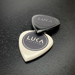 以下が含まれることがあります： 白と黒の2枚のギターピック。どちらもLUKA PLECTRUMSという文字が入った円形のロゴが特徴です。白いピックが手前にあり、黒いピックがその後ろに少し重なるように配置されており、どちらも滑らかで光沢のある仕上がりになっています。