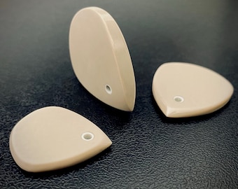 May include: Three smooth, beige, teardrop-shaped guitar picks with a glossy finish. Each pick features a small, circular hole near the pointed end, reinforced with a white ring. The picks are arranged with one standing upright and two lying flat.