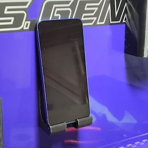 May include: A blue smartphone is mounted on a black holder attached to a purple surface. The surface has the text "RUB" and "J.S. GEM" in white. Other black storage containers hold tools and accessories.