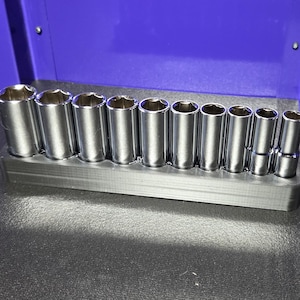 May include: A set of shiny, chrome sockets of varying sizes, neatly arranged in a gray holder. The sockets are cylindrical with hexagonal openings. The holder is rectangular and sits on a dark surface, with a blue background.