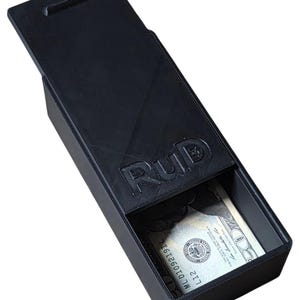 May include: A black rectangular box with a hinged lid, partially open to reveal a twenty-dollar bill. The box has a textured surface and the word "RUD" is embossed on the top. The lid has a small rectangular cutout.