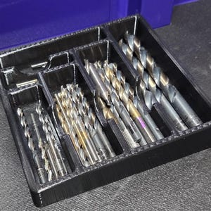 May include: A black plastic drill bit organiser filled with various drill bits. The bits are silver and gold in colour, and are arranged in separate compartments within the organiser. The organiser is on a dark surface.