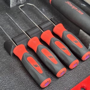 May include: A set of four red and black Snap-on pick tools with curved metal tips. The tools are arranged in a black holder, with the Snap-on logo visible on the handles. A larger Snap-on tool is in the background.