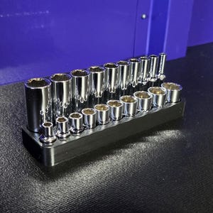 May include: A set of chrome sockets of various sizes, arranged on a black holder. The sockets are cylindrical with a hexagonal opening. The holder is rectangular and sits on a dark surface, with a blue background.